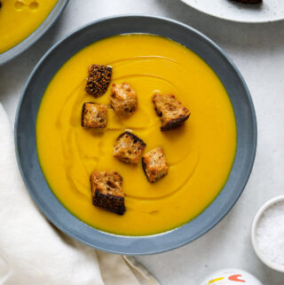 A blue bowl filled with orange, pureed soup, toasted croutons and a drizzle of olive oil.