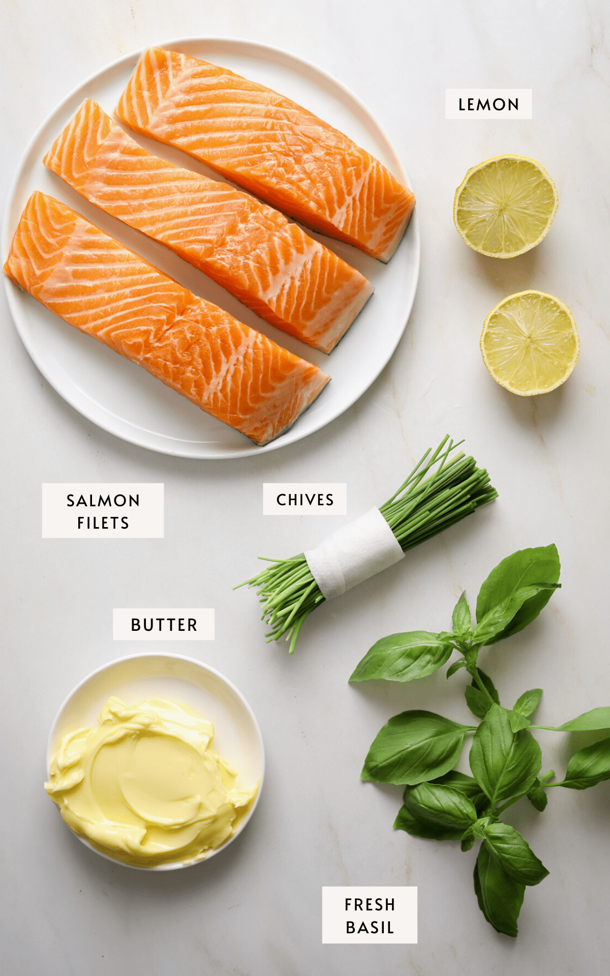 A white plate with three raw salmon filets, a halved lemon, a bundle of chives, a pile of fresh basil leaves and a small white plate of butter.