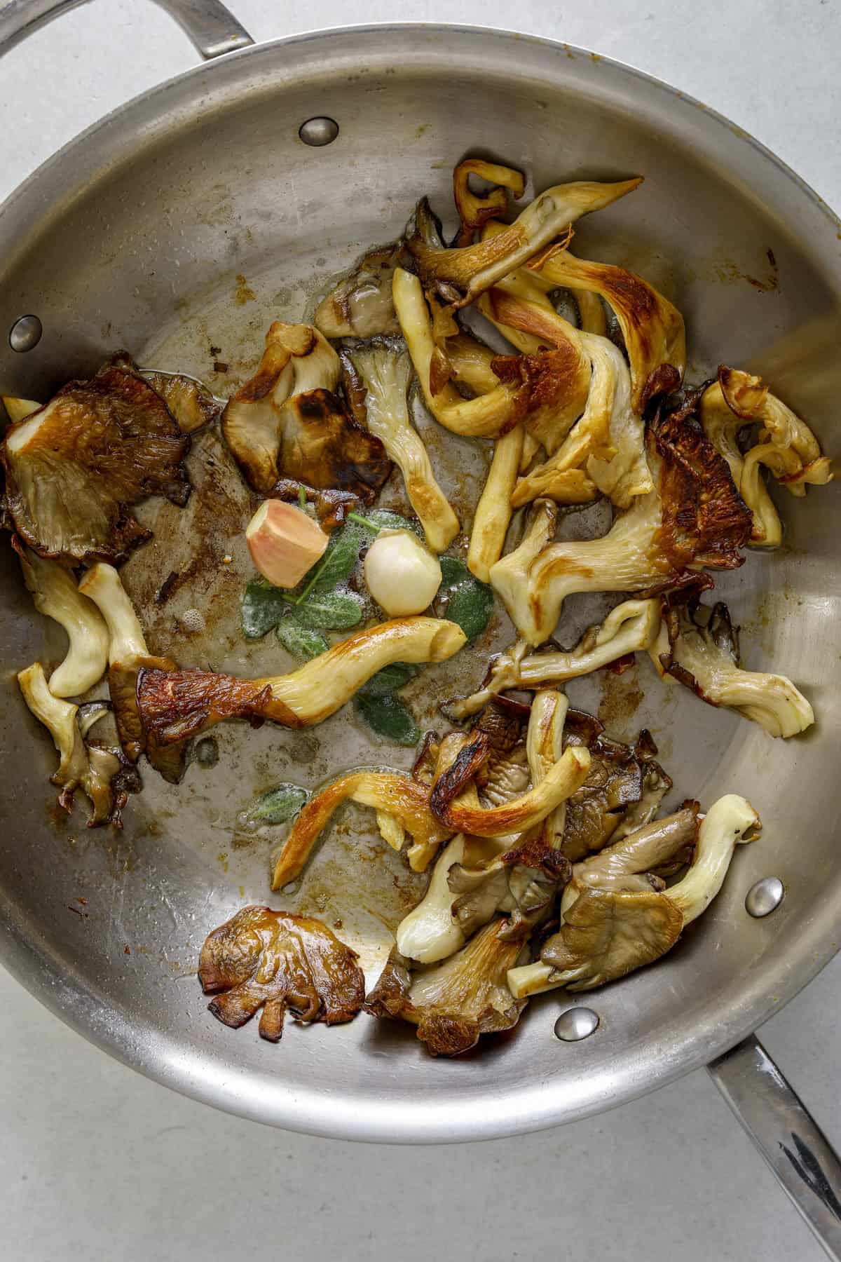 A stainless steel sauté pan filled with mushrooms, garlic cloves and sage leaves.