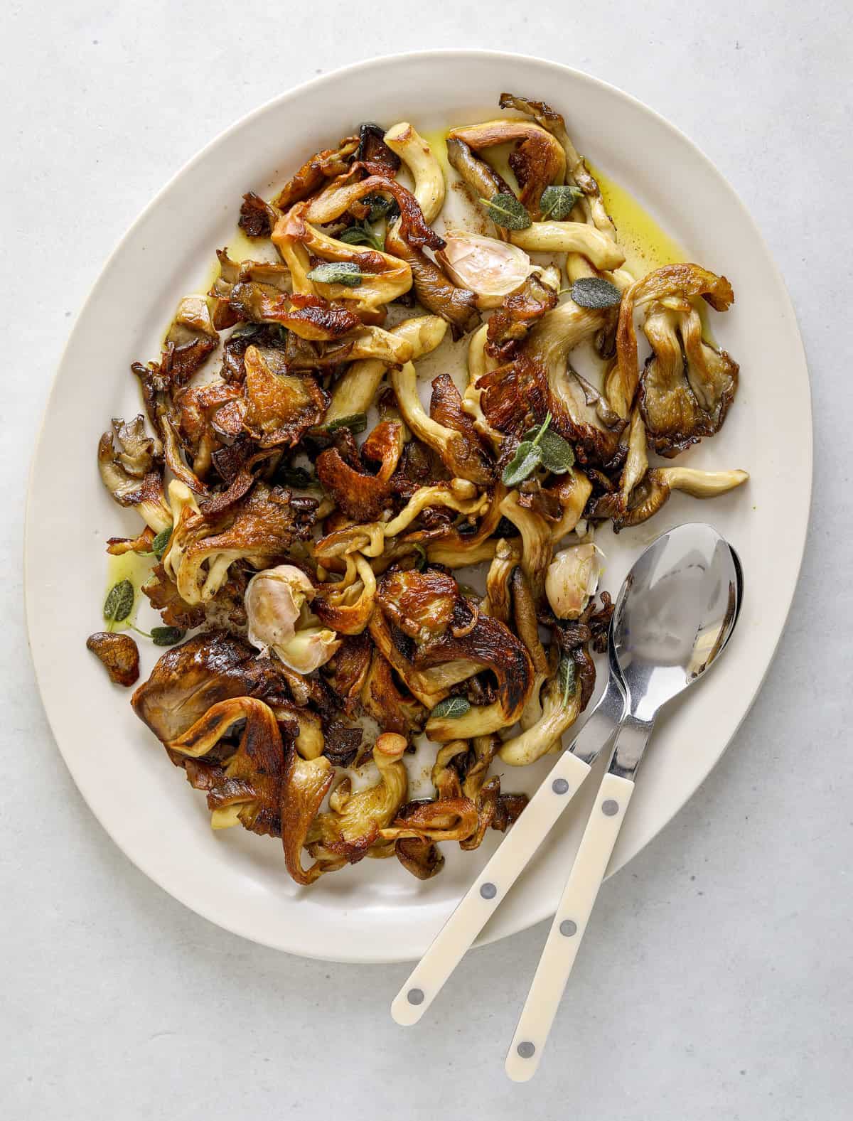 A white oval platter filled with golden-brown roasted oyster mushrooms and crispy sage leaves, two white handled serving spoons lay on the side of the platter.