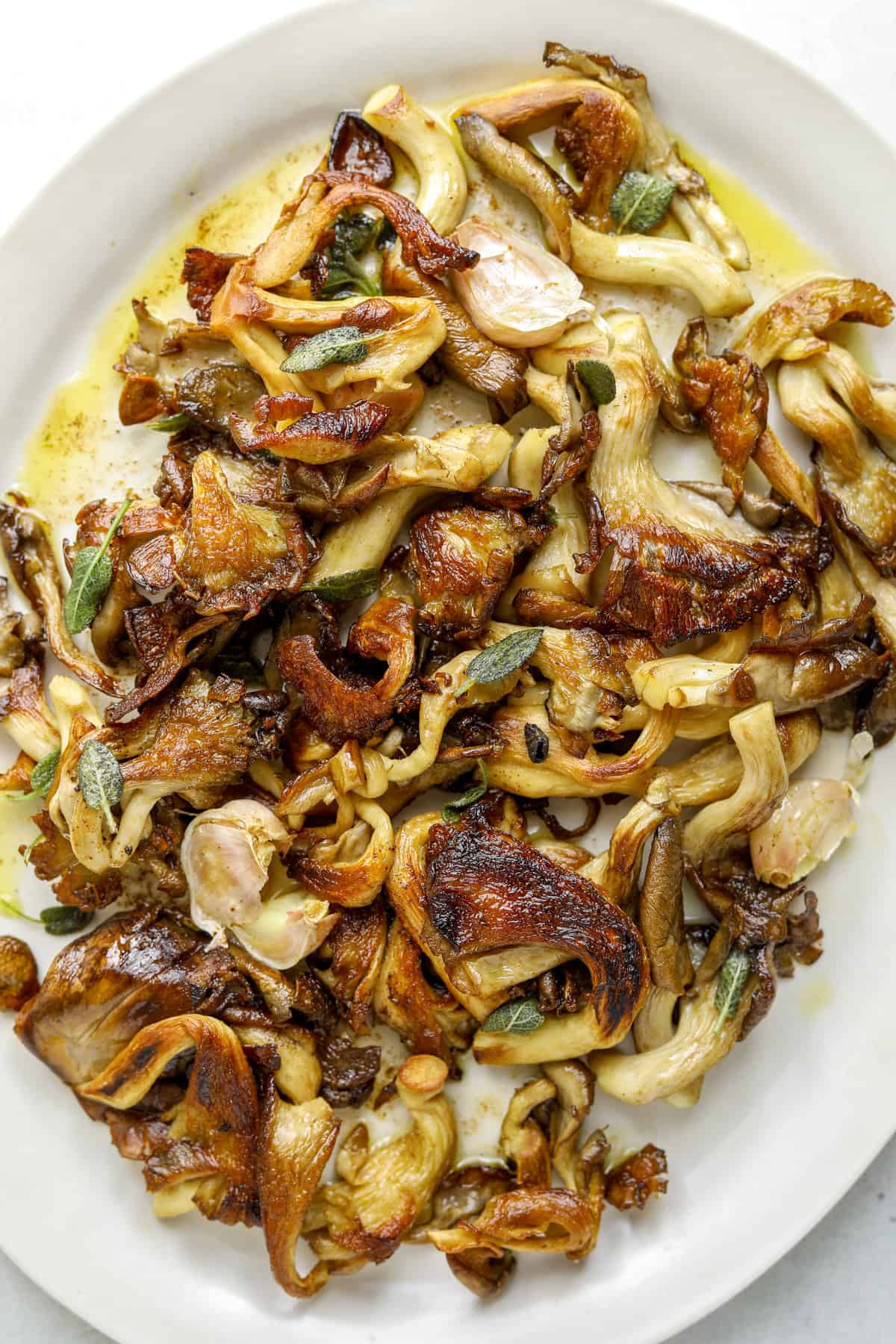 A white oval serving platter piled high with golden-brown roasted oyster mushrooms, crispy sage leaves, whole garlic cloves and pools of nutty brown butter.