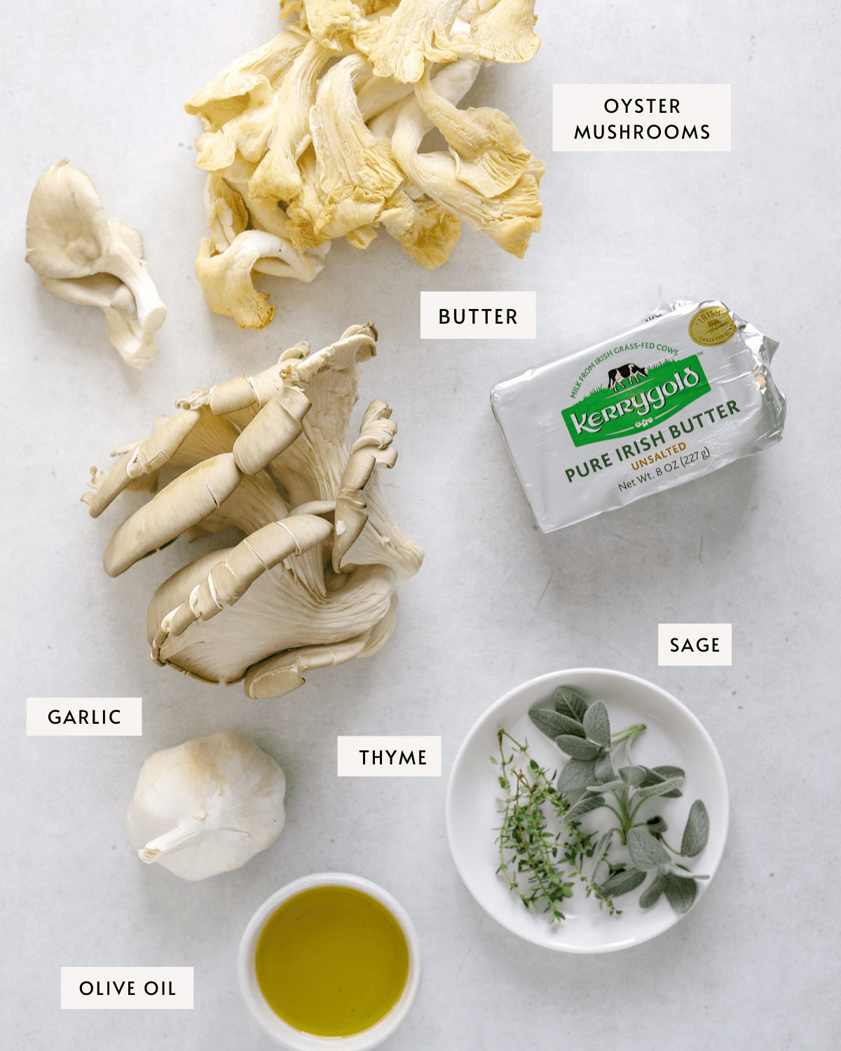 Two clusters of pale gray and light yellow oyster mushrooms, a pack of Irish butter, fresh sage and thyme leaves on a small white dish, a small bowl of olive oil and a bulb of garlic on a light blue/gray background.