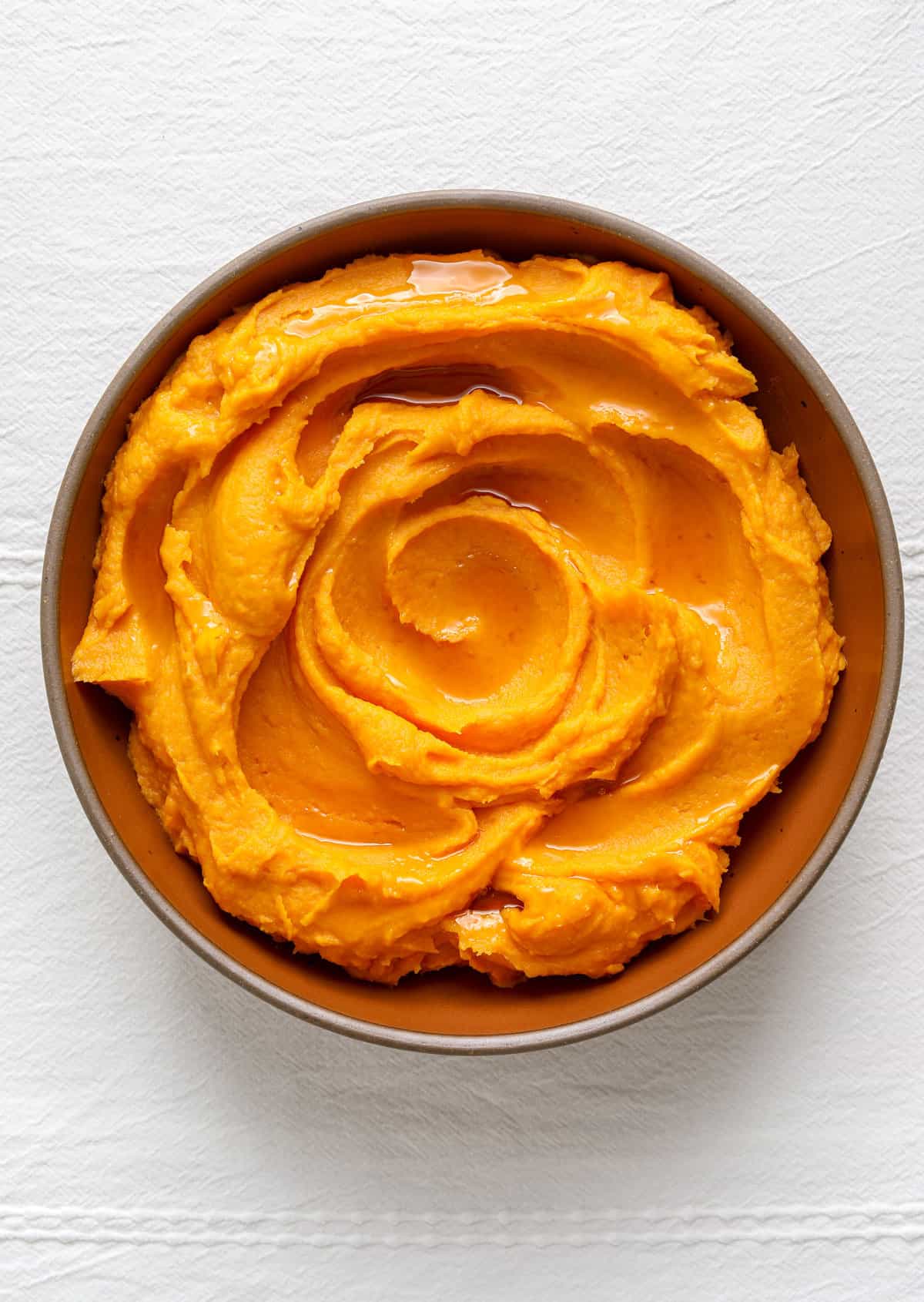 An orange serving bowl filled with sweet potatoes on a white linen table cloth.
