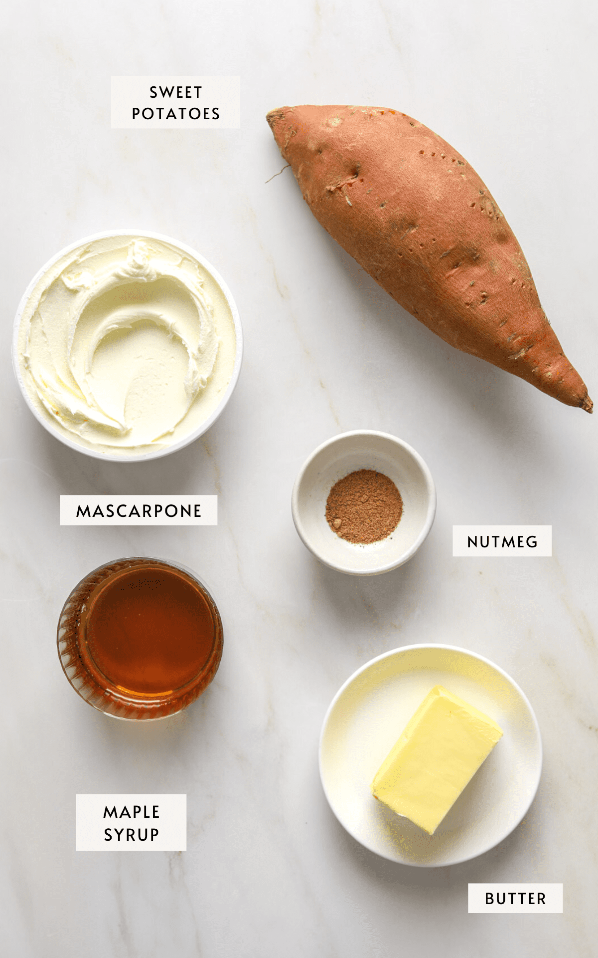 A marble table top with one sweet potato, a container of mascarpone cheese, a small glass dish of maple syrup, a stick of butter and a small dish of ground nutmeg.