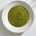 A white ceramic bowl filled with a coarsely puréed green and yellow pesto on a light gray shadowy background.