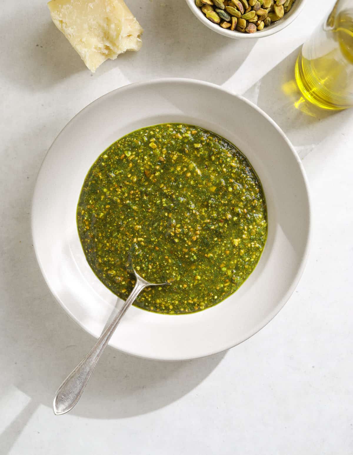 A bowl of green pesto with a silver spoon, surrounded by a bottle of olive oil, a small bowl of pistachios and a hunk of parmesan cheese.