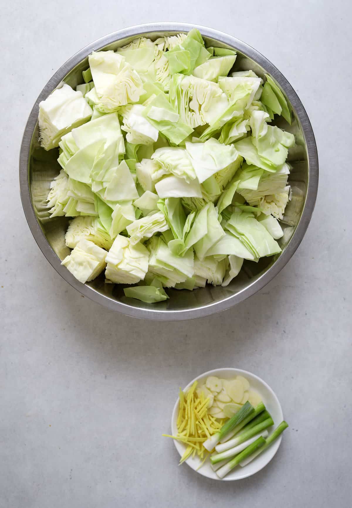 A mixing bowl with chopped cabbage and a small plate with slivered garlic, ginger and green onion.