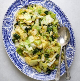 A blue and white platter filled with chopped, sautéed cabbage, bits of ginger and sesame seeds as well as two large silver serving utensils.
