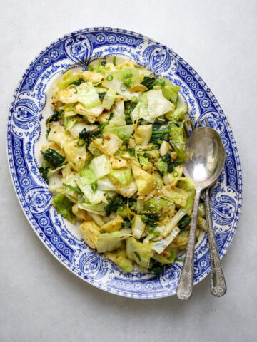 A blue and white platter filled with chopped, sautéed cabbage, bits of ginger and sesame seeds as well as two large silver serving utensils.
