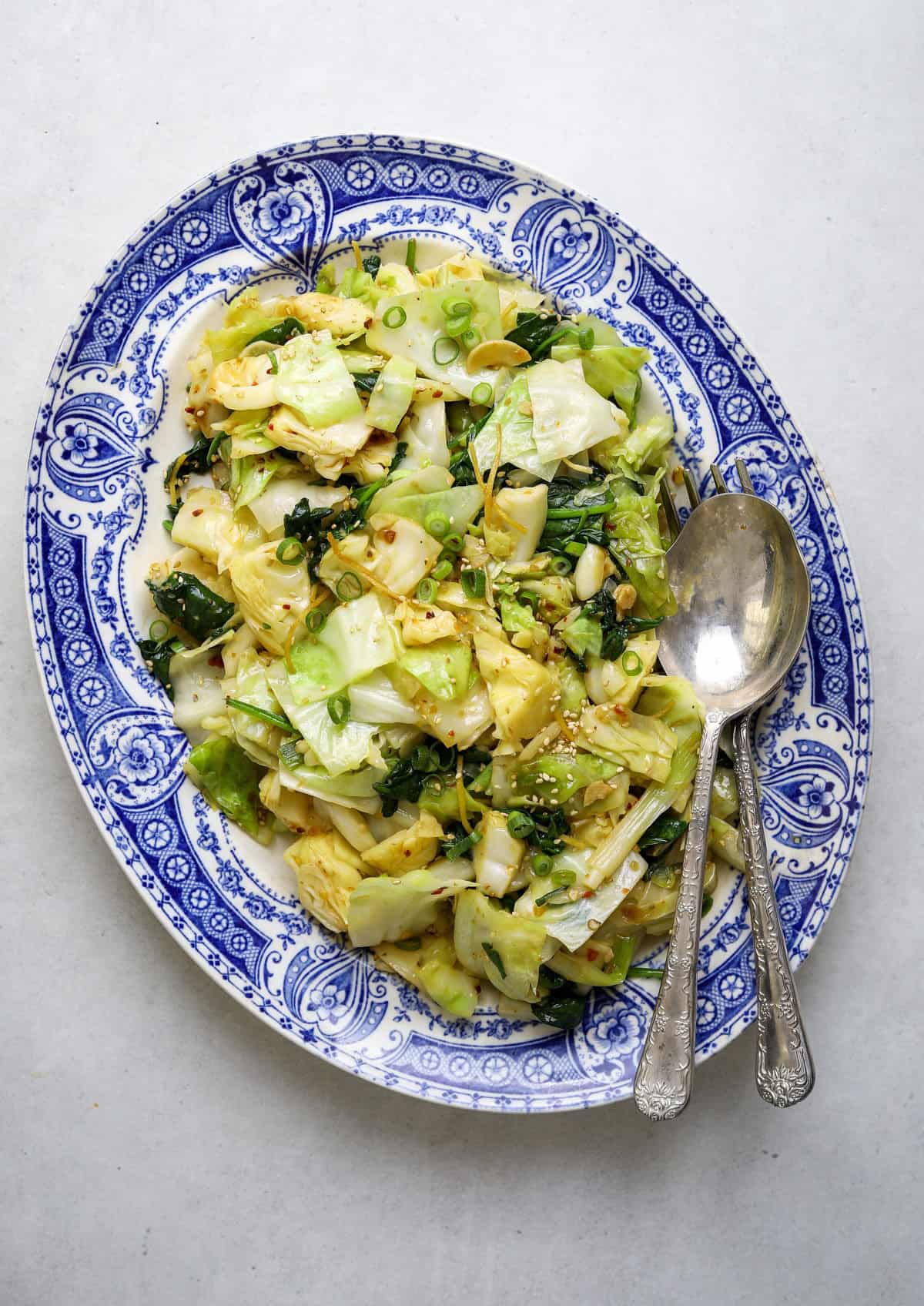 A blue and white platter filled with chopped, sautéed cabbage, bits of ginger and sesame seeds as well as two large silver serving utensils.