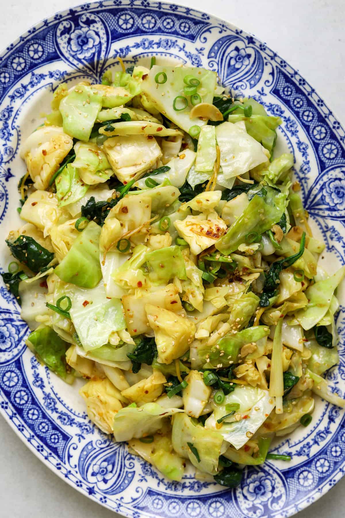 A close up image of pale green and light yellow cooked cabbage with ginger, green onion and sesame seeds on a blue and white platter.