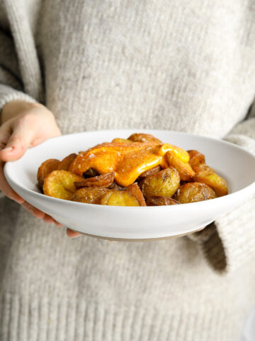 A person in a gray sweater holding a bowl of crispy roasted potatoes with a red sauce.