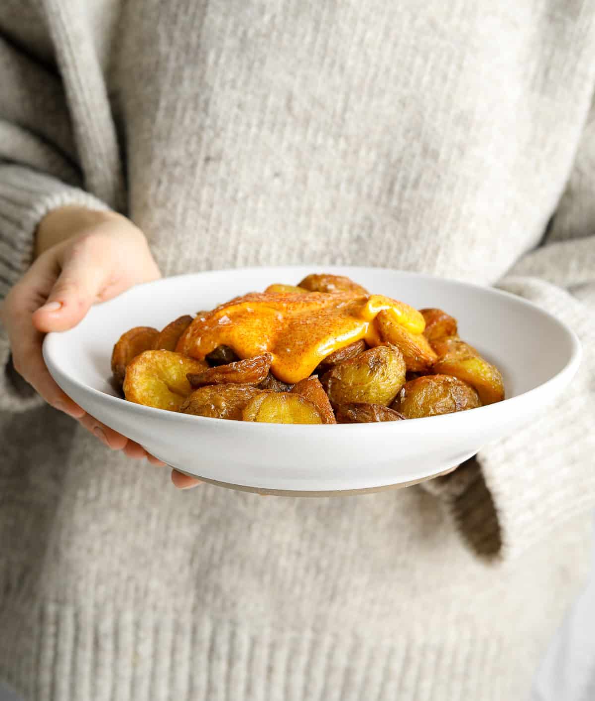 A person in a gray sweater holding a bowl of crispy roasted potatoes with a red sauce.