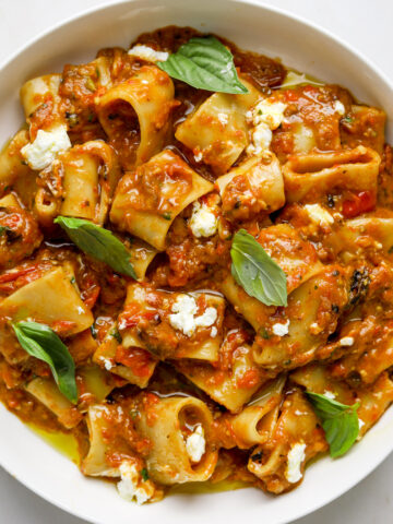 A white bowl filled with pasta tossed in a a roasted tomato eggplant sauce and topped with fresh basil leaves and crumbled goat cheese.