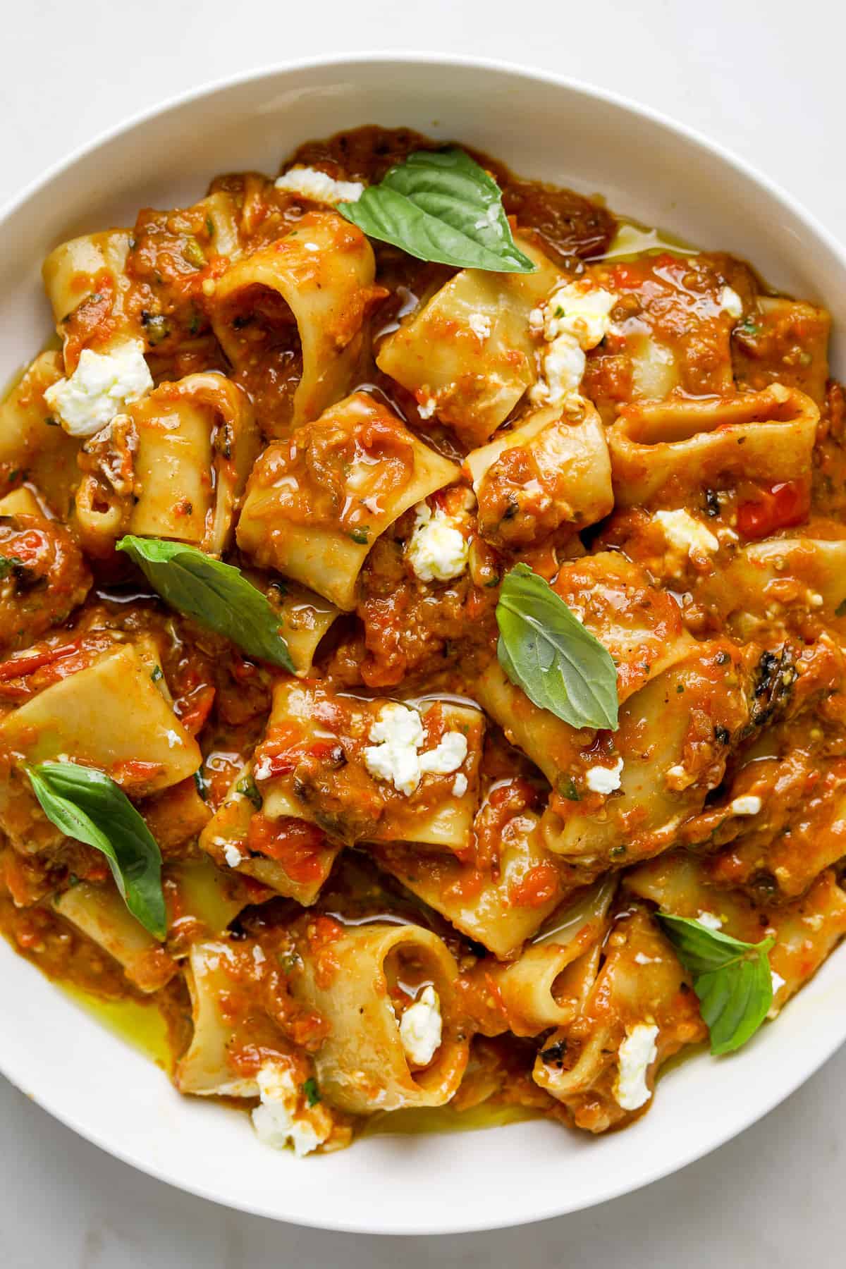 A white bowl filled with pasta tossed in a a roasted tomato eggplant sauce and topped with fresh basil leaves and crumbled goat cheese.