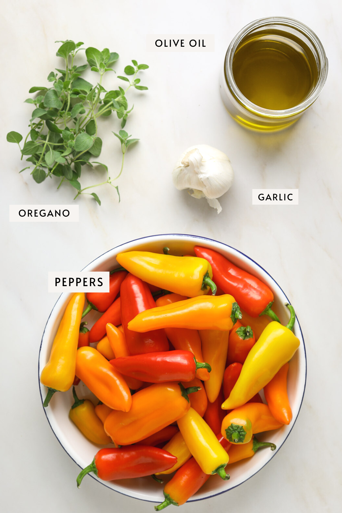 A bowl of baby bell peppers, a bundle of fresh oregano, a jar of olive oil and a bulb of garlic on a marble table top.