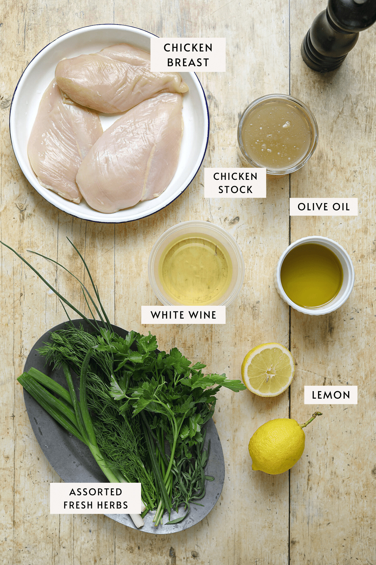 Recipe ingredients measured out individually; a bowl of raw chicken, a white dish of olive oil, a lemon, bundles of fresh herbs and a cup of white wine.