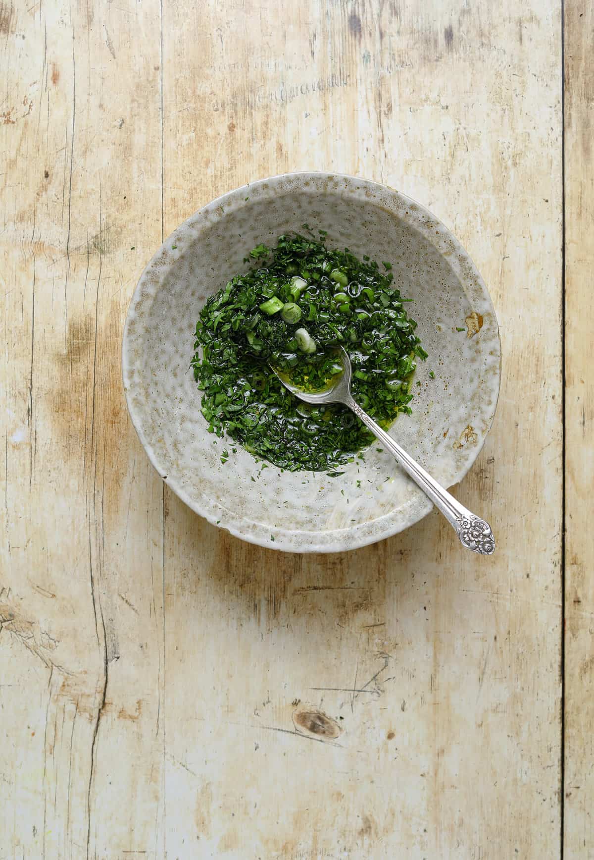 A small, grey, ceramic bowl filled with minced herbs and olive oil on a wooden table top.