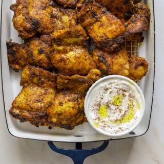 A blue and white enamel Staub baking dish filled with charred chicken thighs and a small dish round of white sauce.