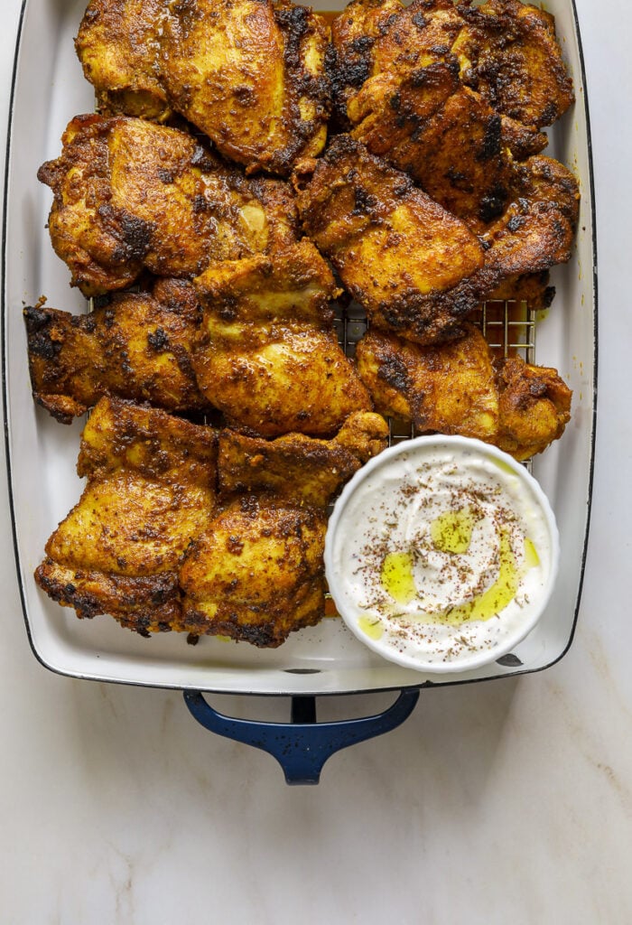 A blue and white enamel Staub baking dish filled with charred chicken thighs and a small dish round of white sauce.