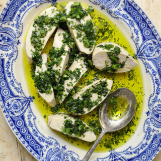 A close up image of slices of poached chicken, drenched in olive oil and chopped herbs, on a blue and white platter with a silver serving spoon.