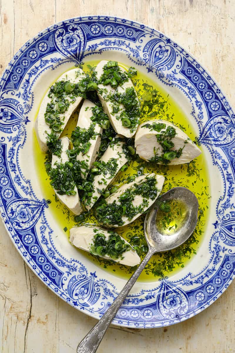 A close up image of slices of poached chicken, drenched in olive oil and chopped herbs, on a blue and white platter with a silver serving spoon.