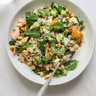 A white ceramic bowl fill with long grain rice, pieces of salmon, fresh mint and garbanzo beans.