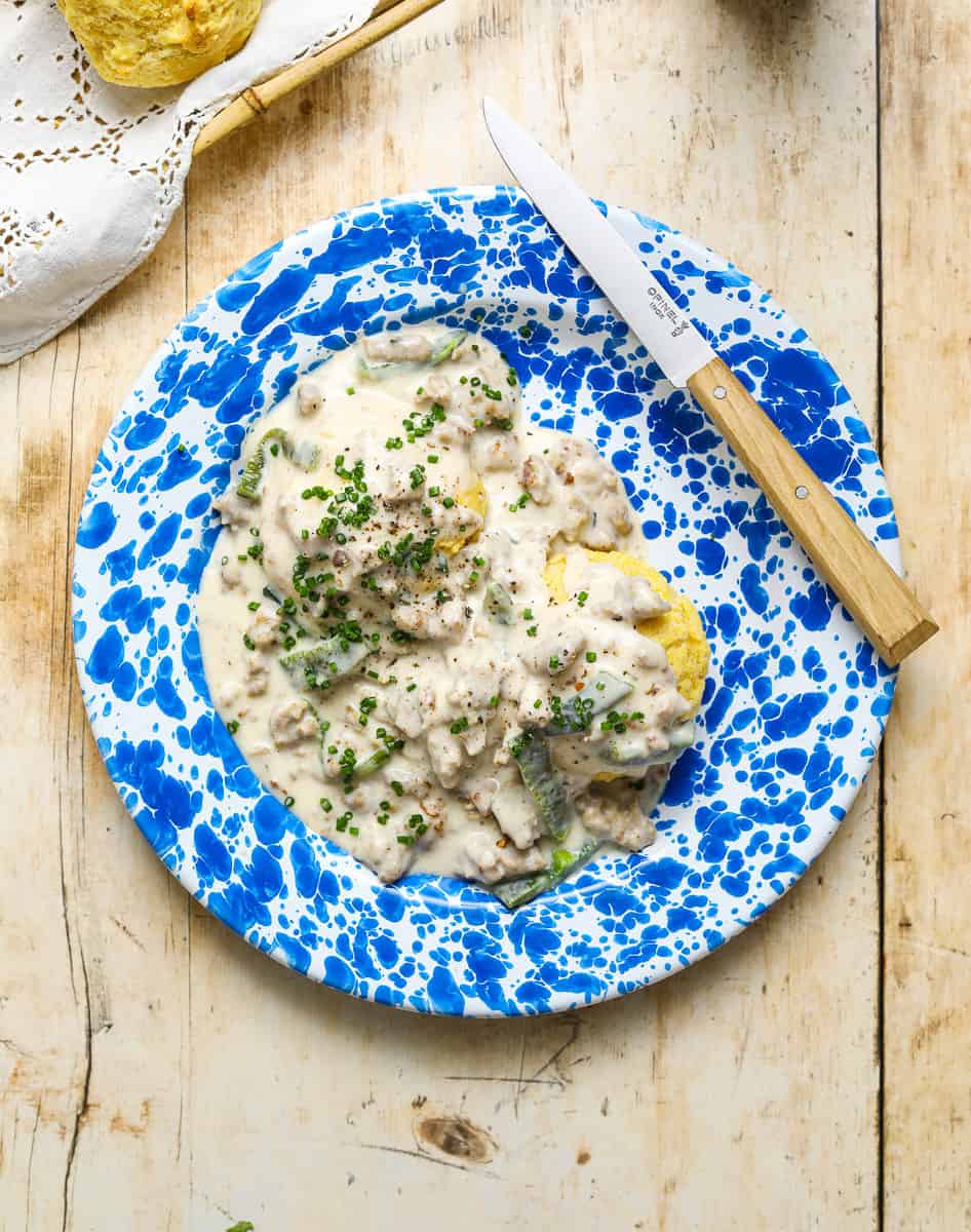 A blue and white enamel plate with yellow corn biscuits and gravy speckled with roasted poblano peppers on a wooden table top.