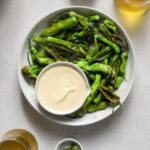 A round platter filled with charred peppers and a dish of creamy dipping sauce surrounded by glasses of beer.