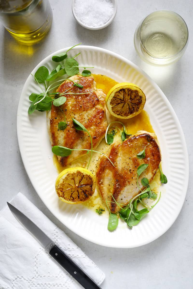 Two seared chicken breasts on a white oval platter topped with watercress and grilled lemons.