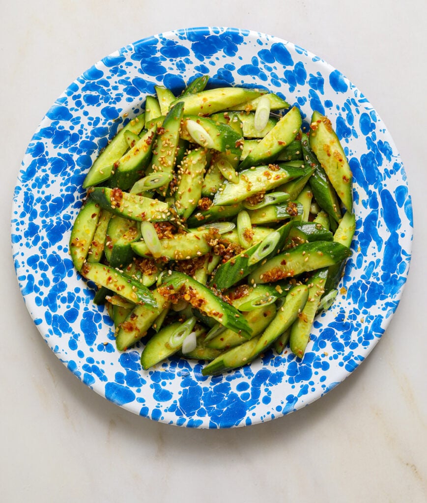 A splattered blue and white enamel plate with cucumber salad on a marble tabletop.