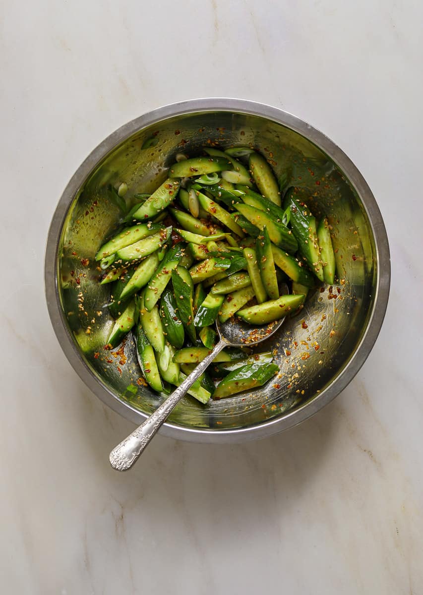A stainless steel mixing bowl filled with sliced cucumbers tossed in chili oil.