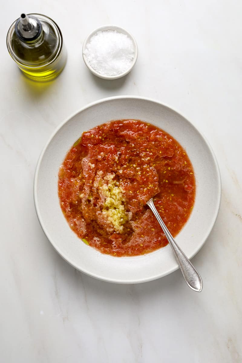 A bowl of tomato puree, grated garlic and olive oil on a marble tabletop.