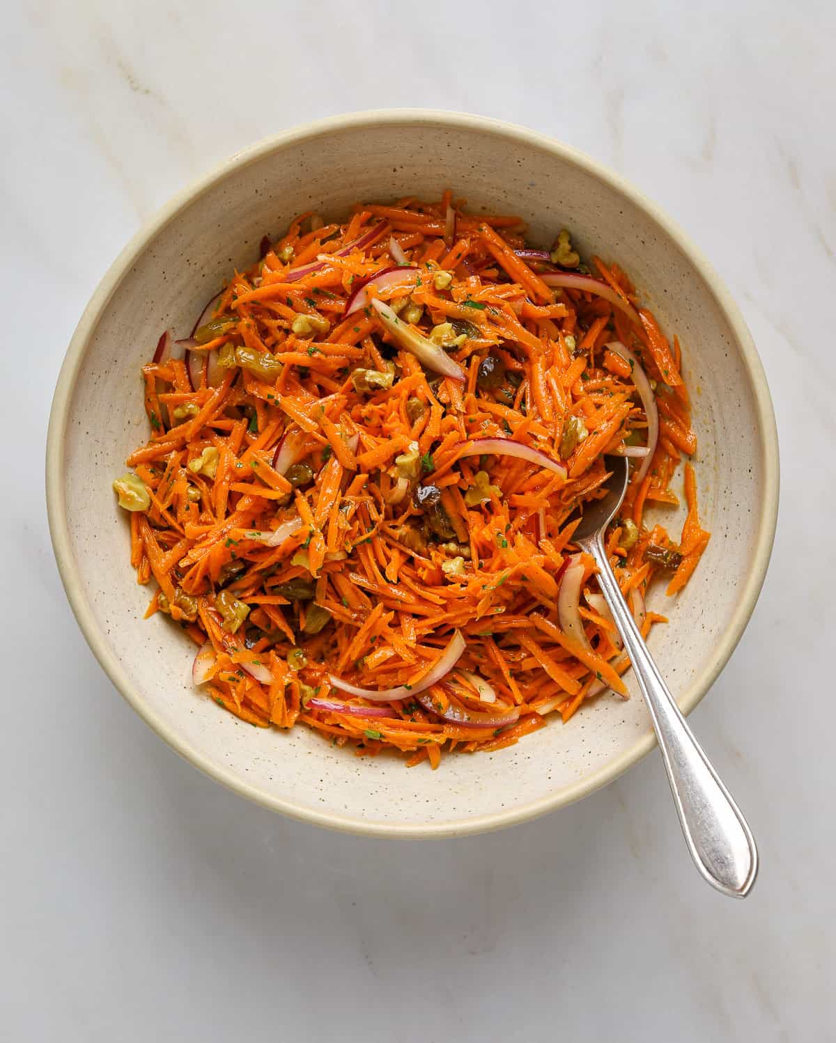 A speckled ceramic bowl filled with a grated carrot slaw, red onion slivers and raisins.