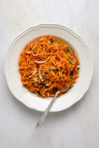 An antique white rimmed bowl filled with carrot salad and a silver spoon on a marble tabletop.