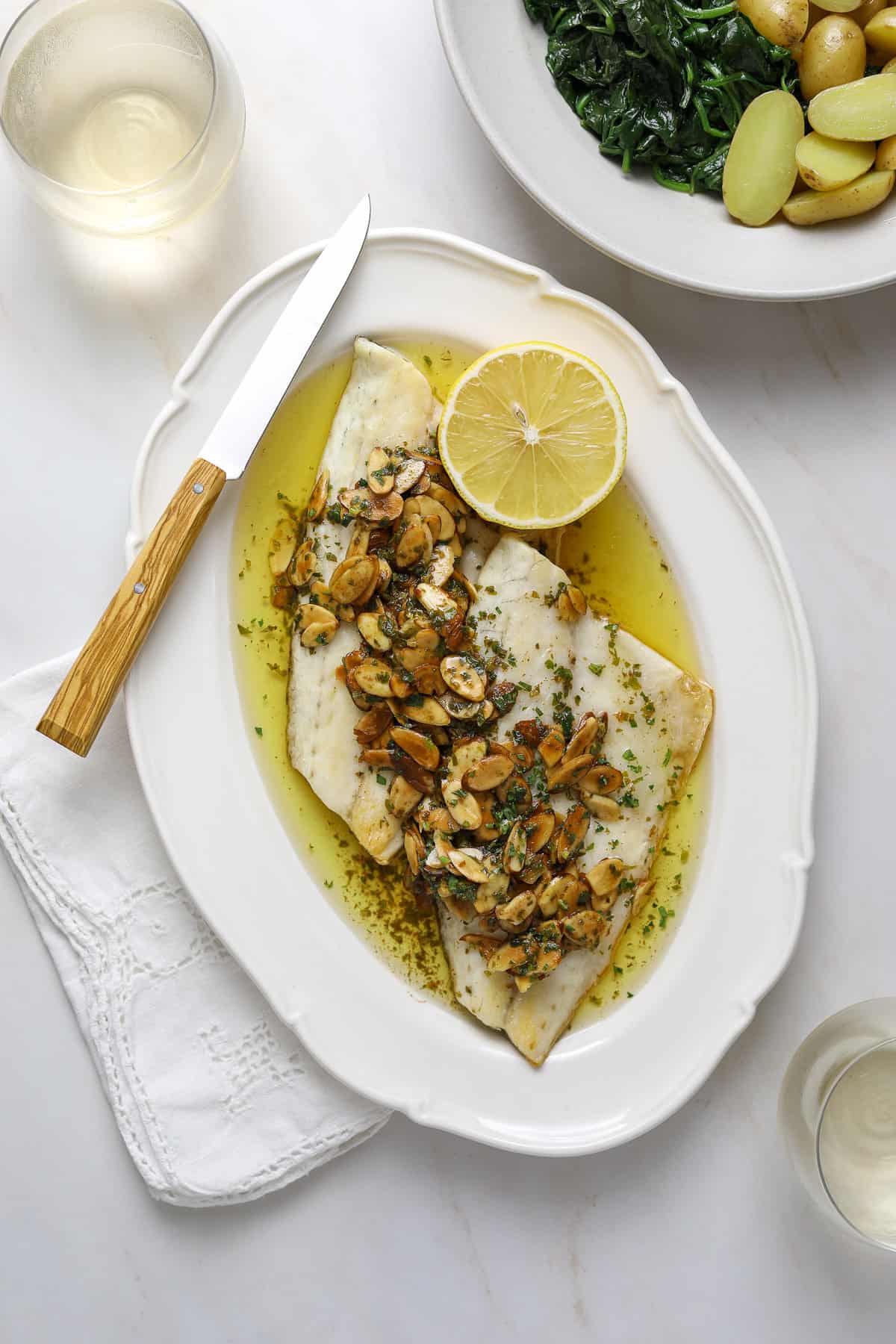 A platter of fish covered in sauce on a marble tabletop with a linen napkin and two glasses of white wine.