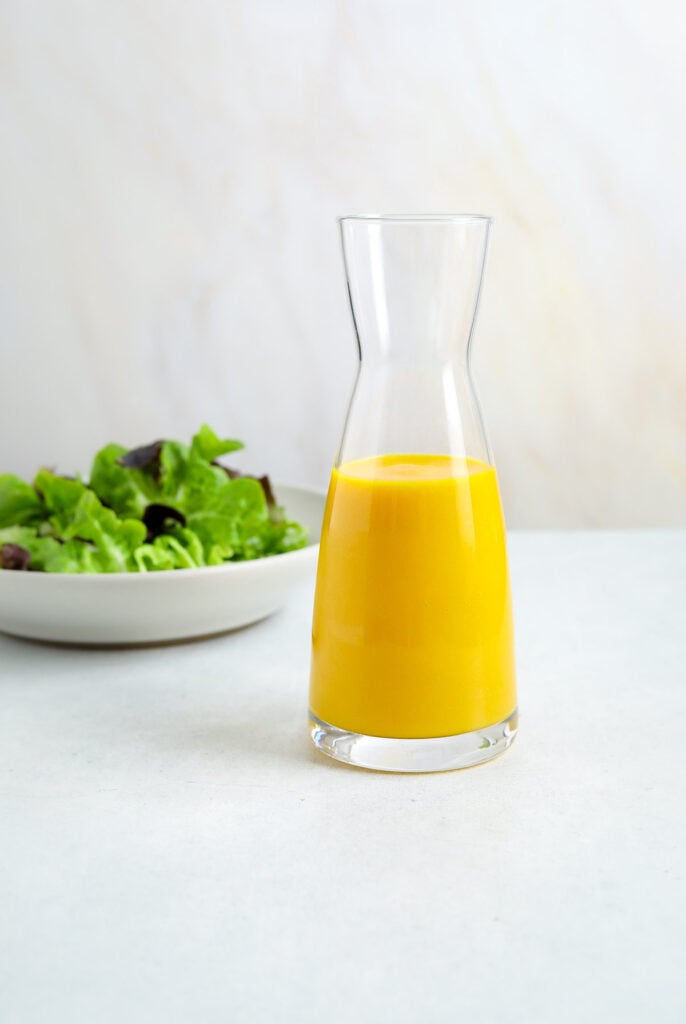 A glass bottle of bright orange salad dressing with a bowl of lettuce in the foreground.