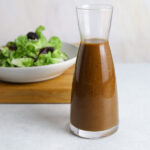 A glass bottle of salad dressing with a bowl of fresh lettuce in the background.