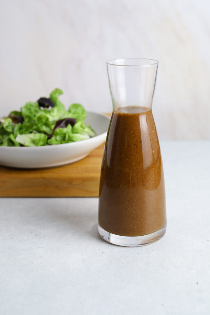 A glass bottle of salad dressing with a bowl of fresh lettuce in the background.