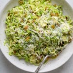 A white speckled ceramic bowl filled with thinly sliced brussels sprouts salad, grated cheese and a silver spoon.