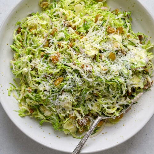 A white speckled ceramic bowl filled with thinly sliced brussels sprouts salad, grated cheese and a silver spoon.