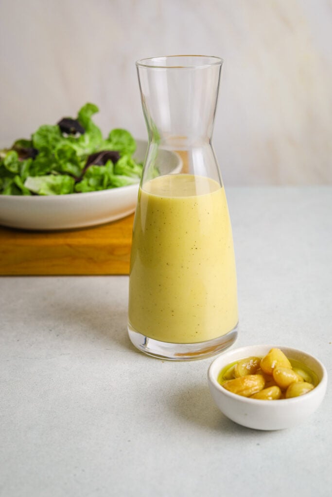 A glass bottle filled with creamy salad dressing. A small dish of roasted garlic cloves in the foreground.