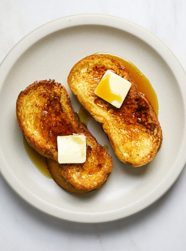 Two pieces of french toast topped with pads of butter and maple syrup on a white, ceramic plate.