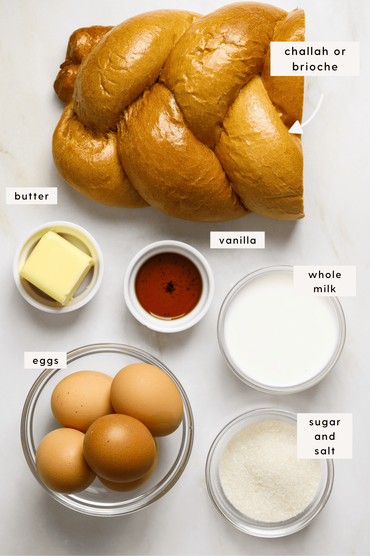 A half loaf of challah, brown eggs in a small glass bowl, sugar, whole milk and vanilla extract.
