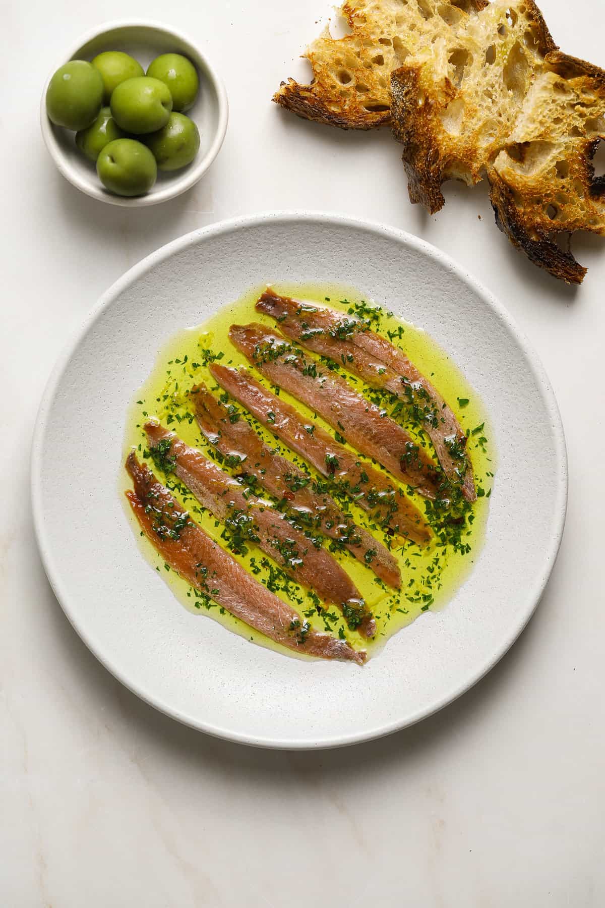 A plate of anchovies drenched in olive oil and herbs with sliced bread on the side and a small bowl of green olives.