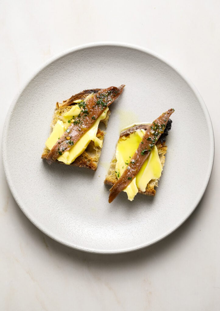 Two crostini topped with butter and and marinated anchovies on a white plate.