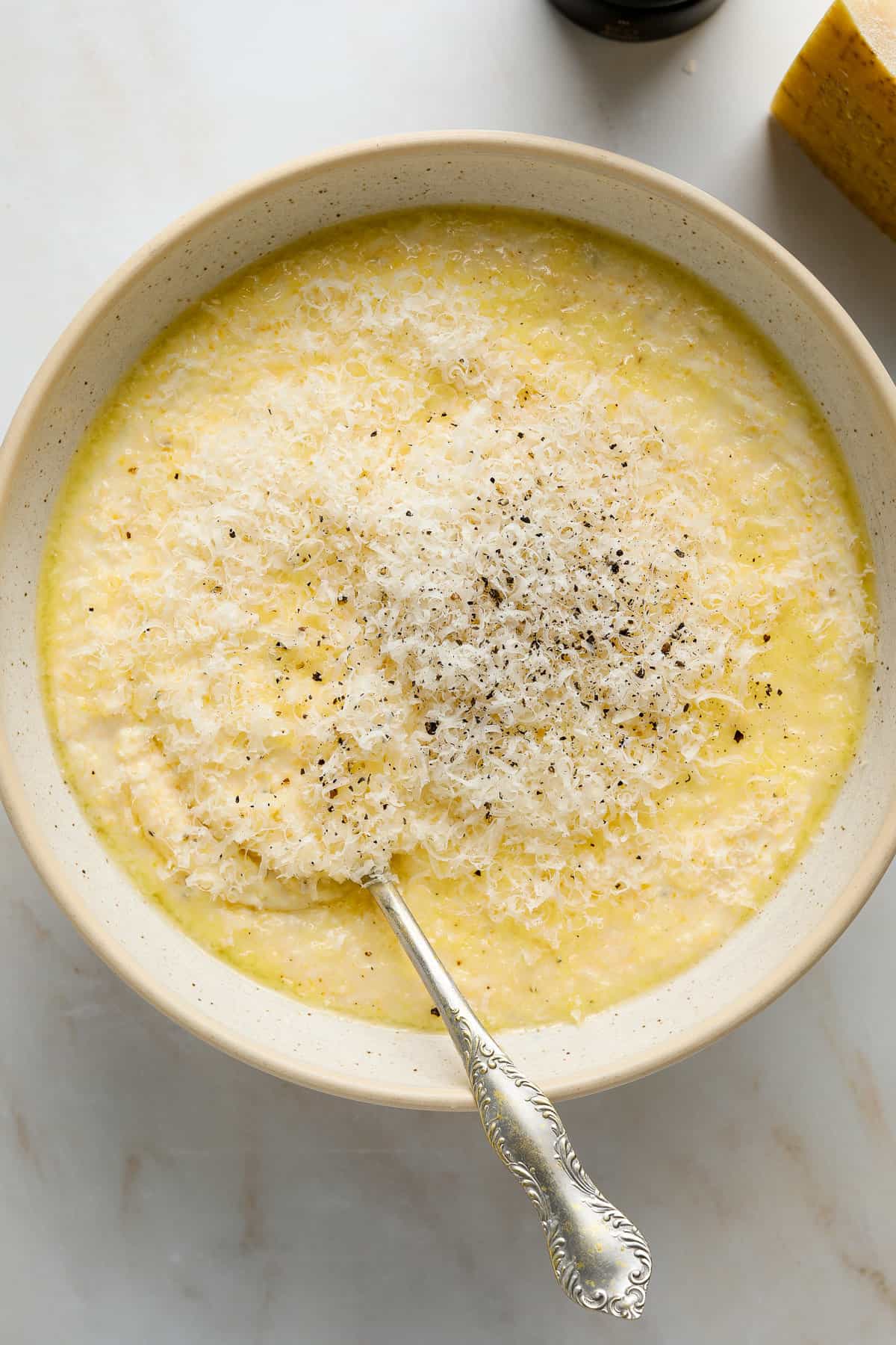 A silver, antique spoon digging into a bowl of creamy, pale yellow polenta topped with grated parmesan cheese and cracked black pepper.