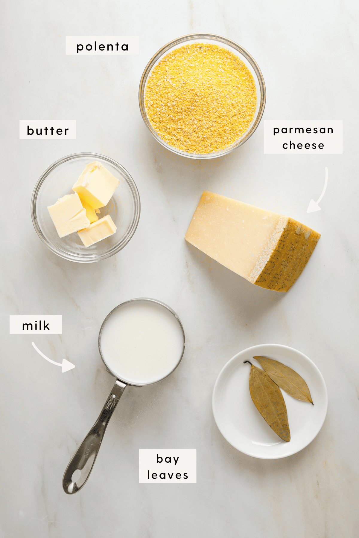 Recipe ingredients individually portioned in small bowls; dried polenta, cubes of butter, a hunk of parmesan cheese, a measuring cup of milk, two bay leaves.