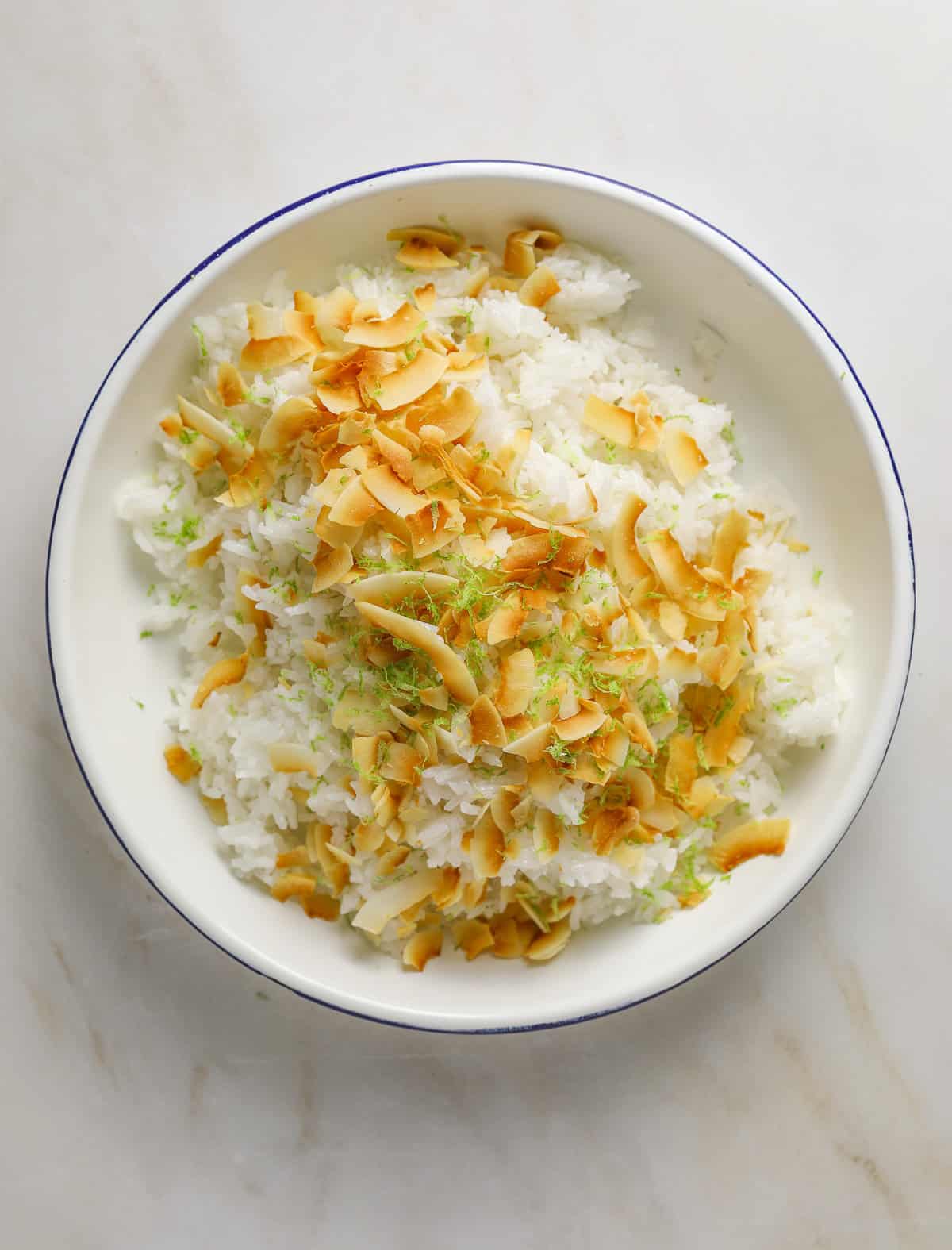 A white and blue enamel bowl filled with steamed rice, toasted coconut flakes and lime zest.