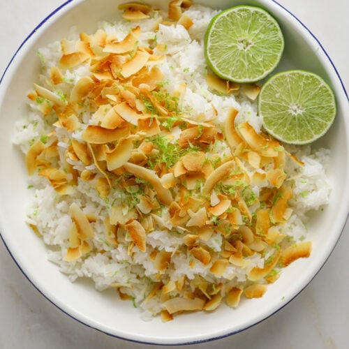 A bowl of rice, coconut and two halves of lime on a marble tabletop.